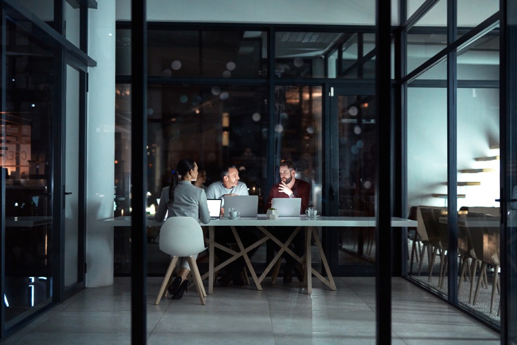 Three People Seated At A Table In A Room With A Glass Wall, Engaged In Conversation.